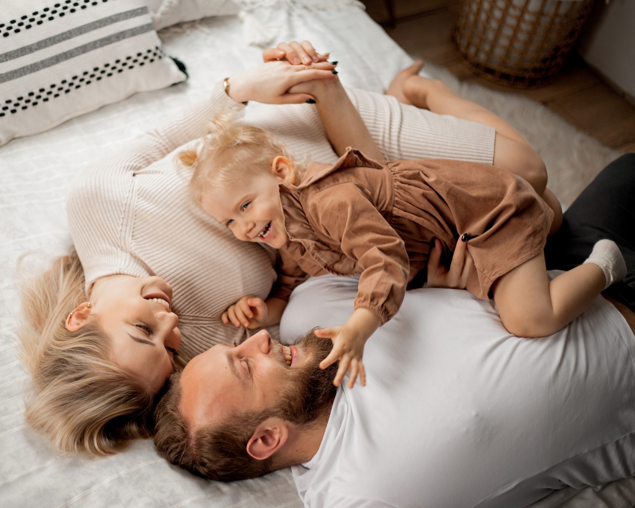 Uśmiechnięta rodzina leży na łóżku: mama w beżowym swetrze, tata w białej koszulce, a mała dziewczynka w brązowej sukience bawi się brodą taty.