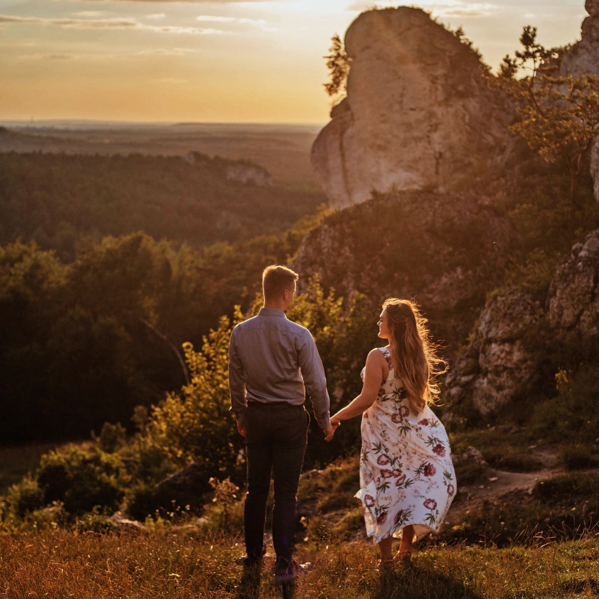 Para trzymająca się za ręce na tle malowniczego krajobrazu z formacjami skalnymi i zachodzącym słońcem, kobieta w kwiecistej sukience, mężczyzna w koszuli i spodniach.
