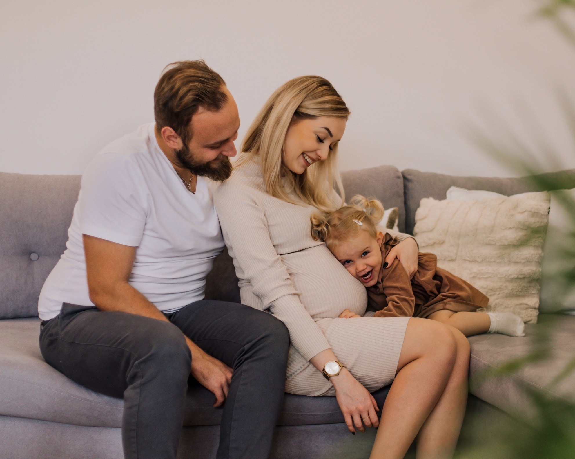 Uśmiechnięta, przyszła mama w beżowej sukience siedzi na szarej kanapie z mężem i córeczką, która przytula się do jej brzucha. Rodzinna fotografia w jasnym wnętrzu.