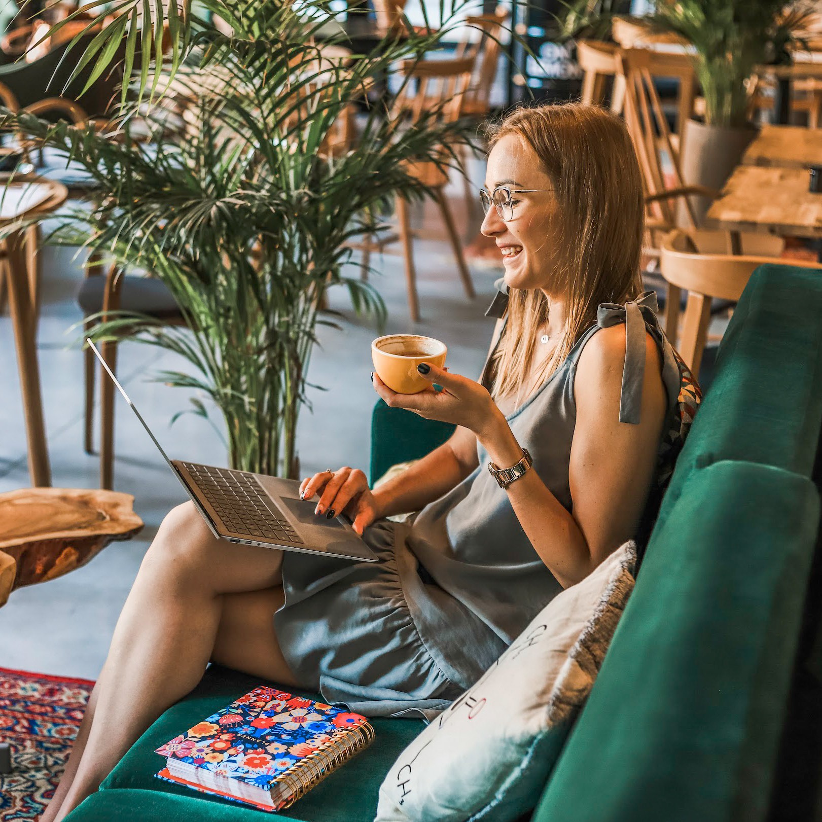 Uśmiechnięta kobieta w okularach siedzi na zielonej kanapie z laptopem i żółtą filiżanką kawy, obok leży kolorowy planer, w tle widoczne rośliny i drewniane krzesła kawiarni.
