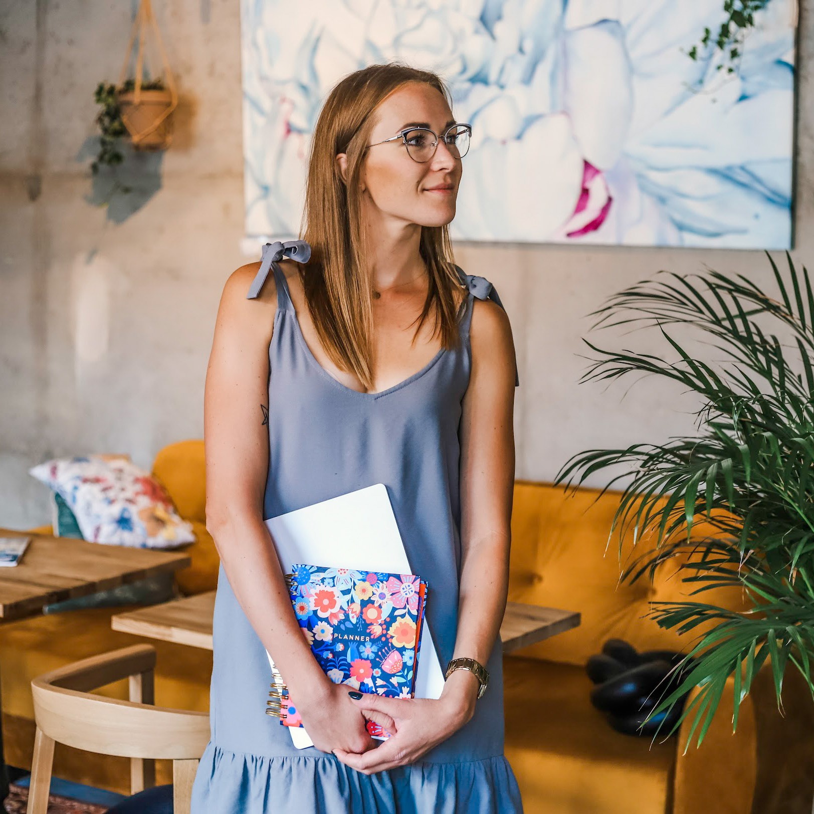 Kobieta w okularach trzymająca notatnik i białą teczkę na tle nowoczesnego wnętrza z żółtą kanapą i obrazem z motywem kwiatowym.