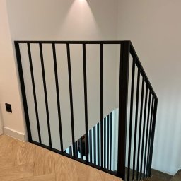 MetalSide - Nowoczesna, czarna balustrada schodowa z pionowymi szczeblami, oświetlona punktowym światłem. Parkiet w jodełkę na podłodze. Minimalistyczny design.
