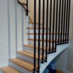 MetalSide - Nowoczesne schody z drewnianymi stopniami i czarną, pionową balustradą. Częściowo szklana barierka na górze. Widok z dołu, oświetlenie naturalne.