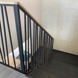 MetalSide - Nowoczesna, czarna balustrada schodowa z drewnianym elementem górnym, widok z góry na ciemne schody i jasną ścianę. Minimalistyczny design.