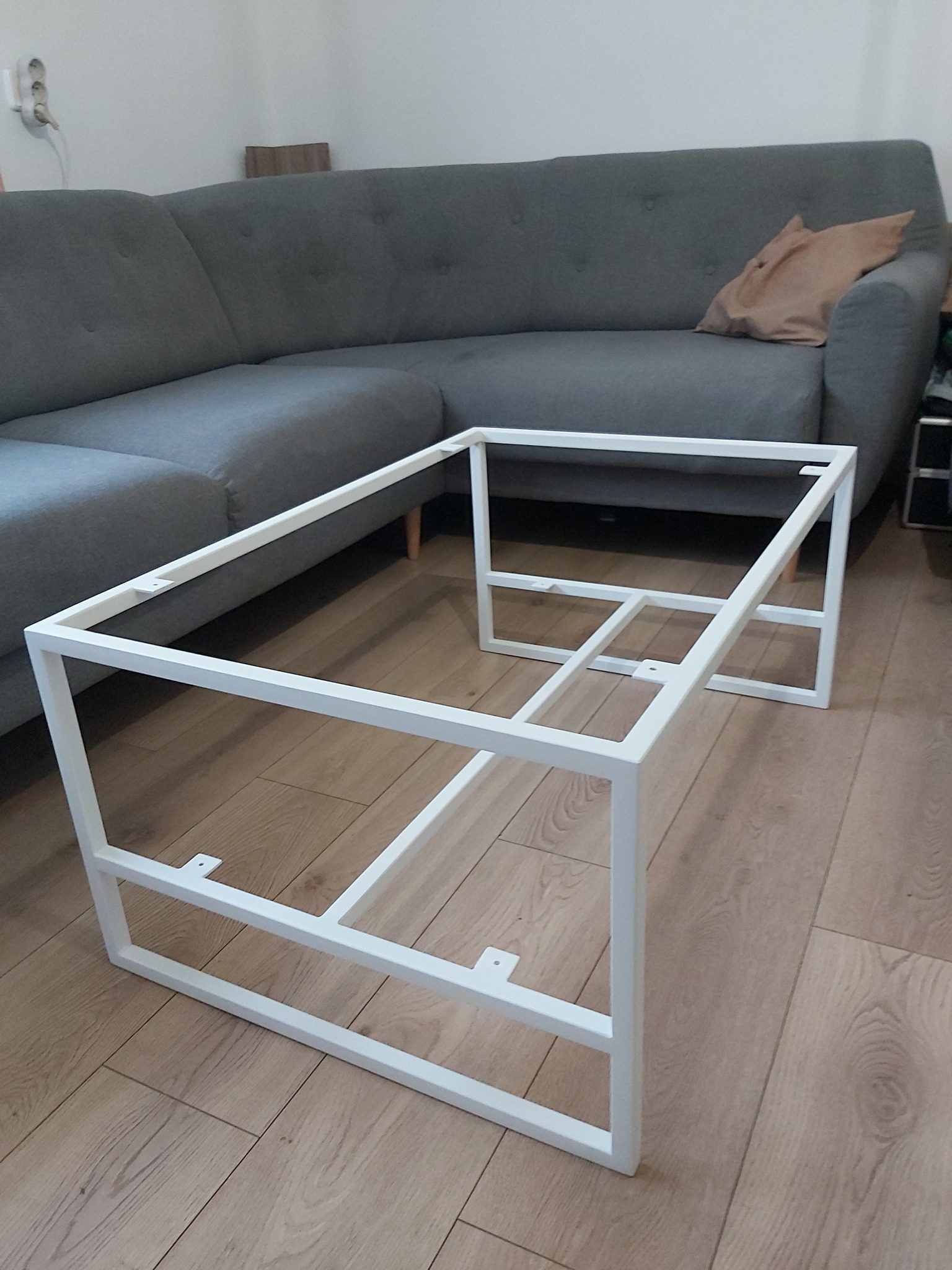 Biała, metalowa rama stolika kawowego, widoczna na tle szarej sofy i drewnianej podłogi.