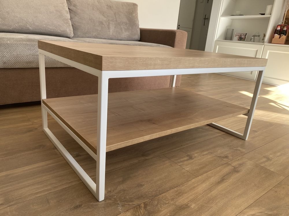 Stolik kawowy z blatem i półką z płyty imitującej jasne drewno, na białym metalowym stelażu, na podłodze z paneli, w tle sofa i zabudowa meblowa.
