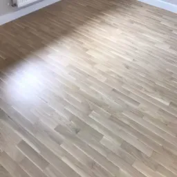 Jasna, wycyklinowana podłoga parkietowa ułożona w jodełkę, widoczny fragment grzejnika i białe listwy przypodłogowe, pomieszczenie po remoncie.