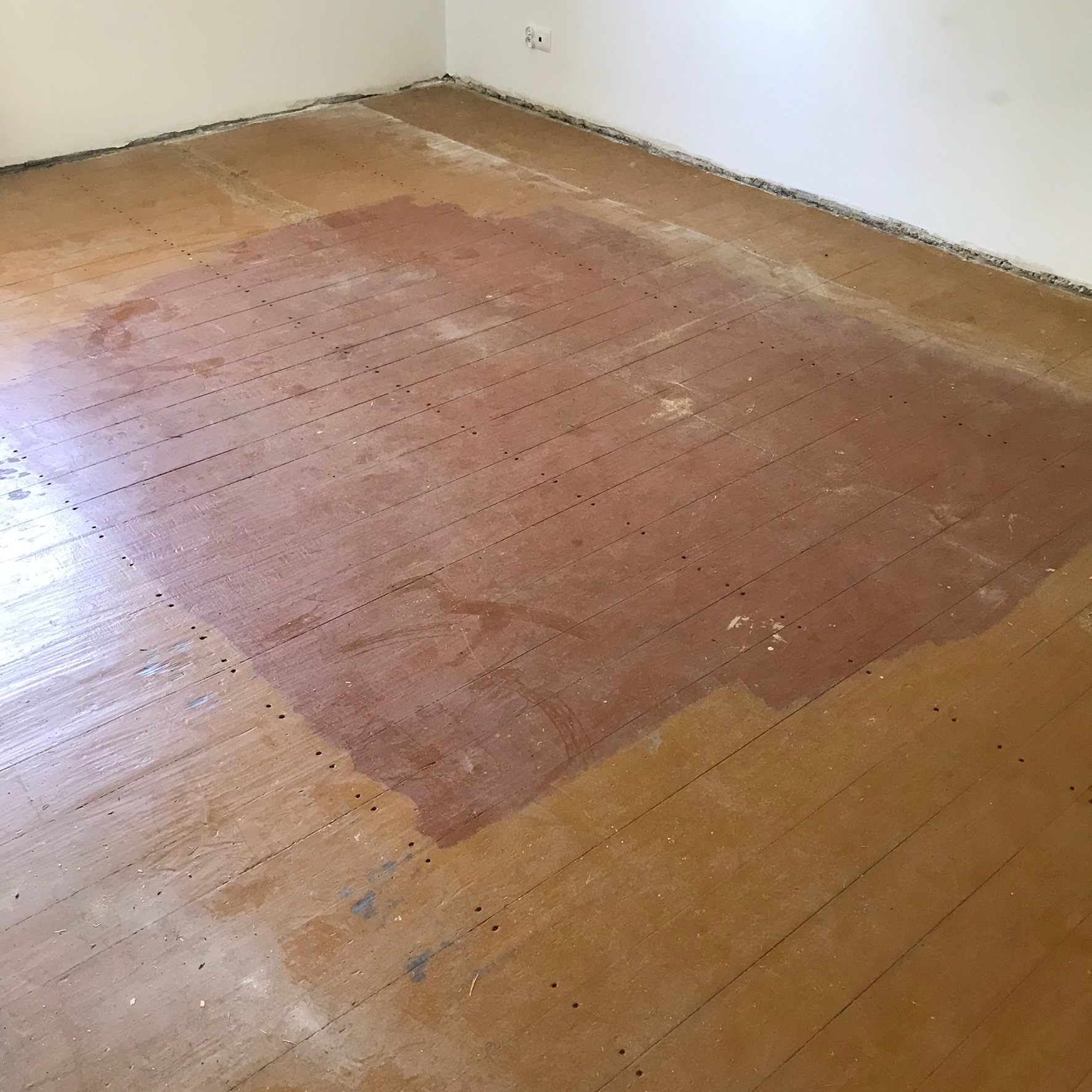 Fragment podłogi z desek, częściowo pokryty świeżą, ciemniejszą warstwą lakieru lub bejcy, ukazujący różnicę w kolorze i strukturze drewna przed i po renowacji.