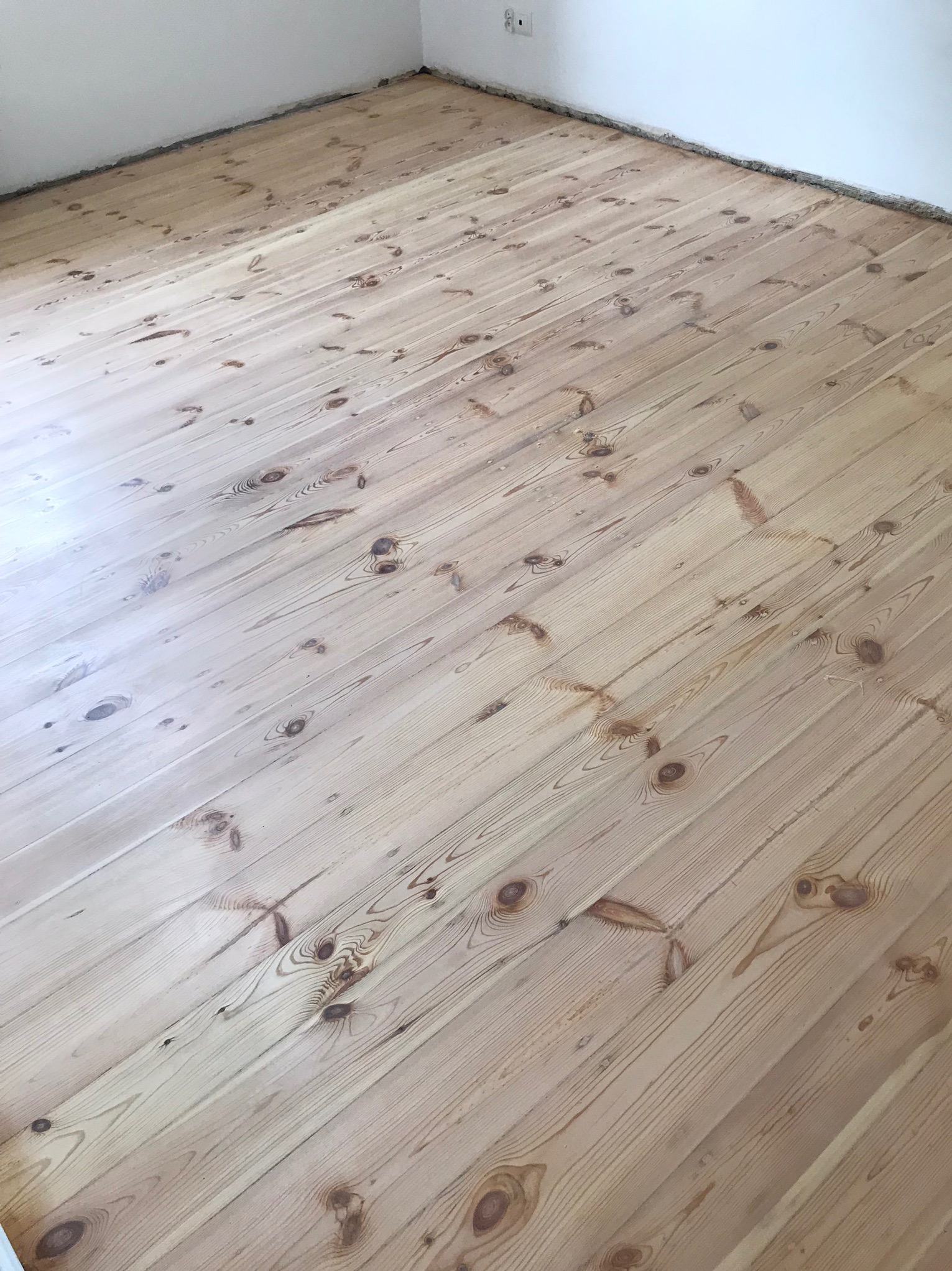 Jasna, świeżo wycyklinowana podłoga z desek sosnowych w pustym pomieszczeniu, widoczne ślady po szlifowaniu przy ścianach.