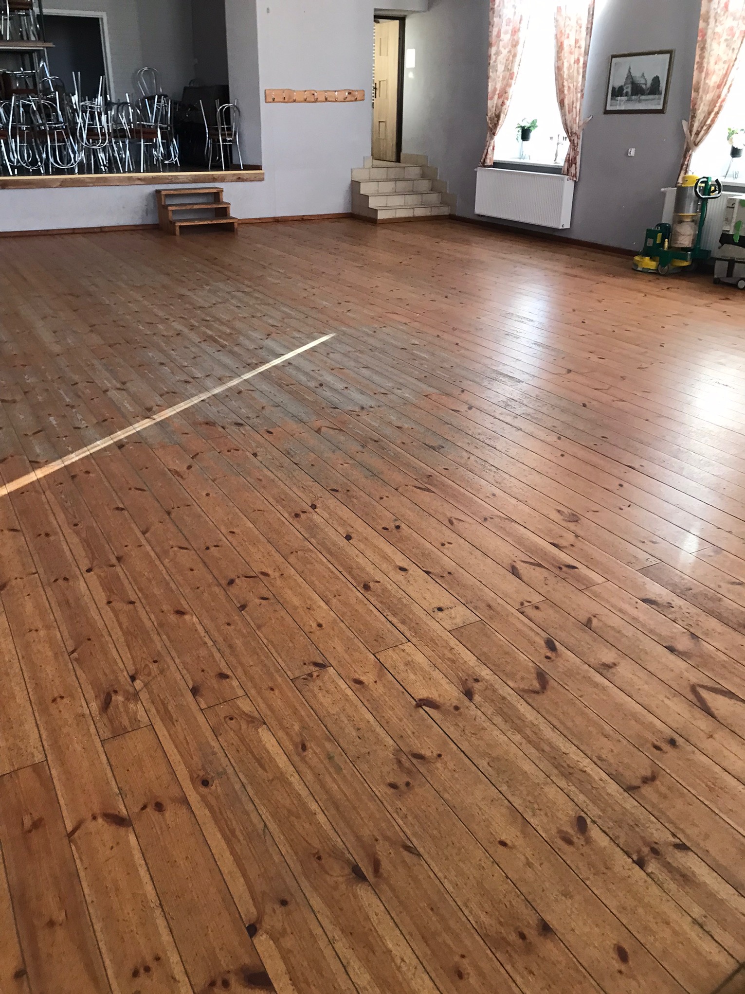 Sala z drewnianą podłogą w trakcie renowacji, widoczna linia oddzielająca obszar już odnowiony od obszaru wymagającego obróbki, w tle krzesła i schody.
