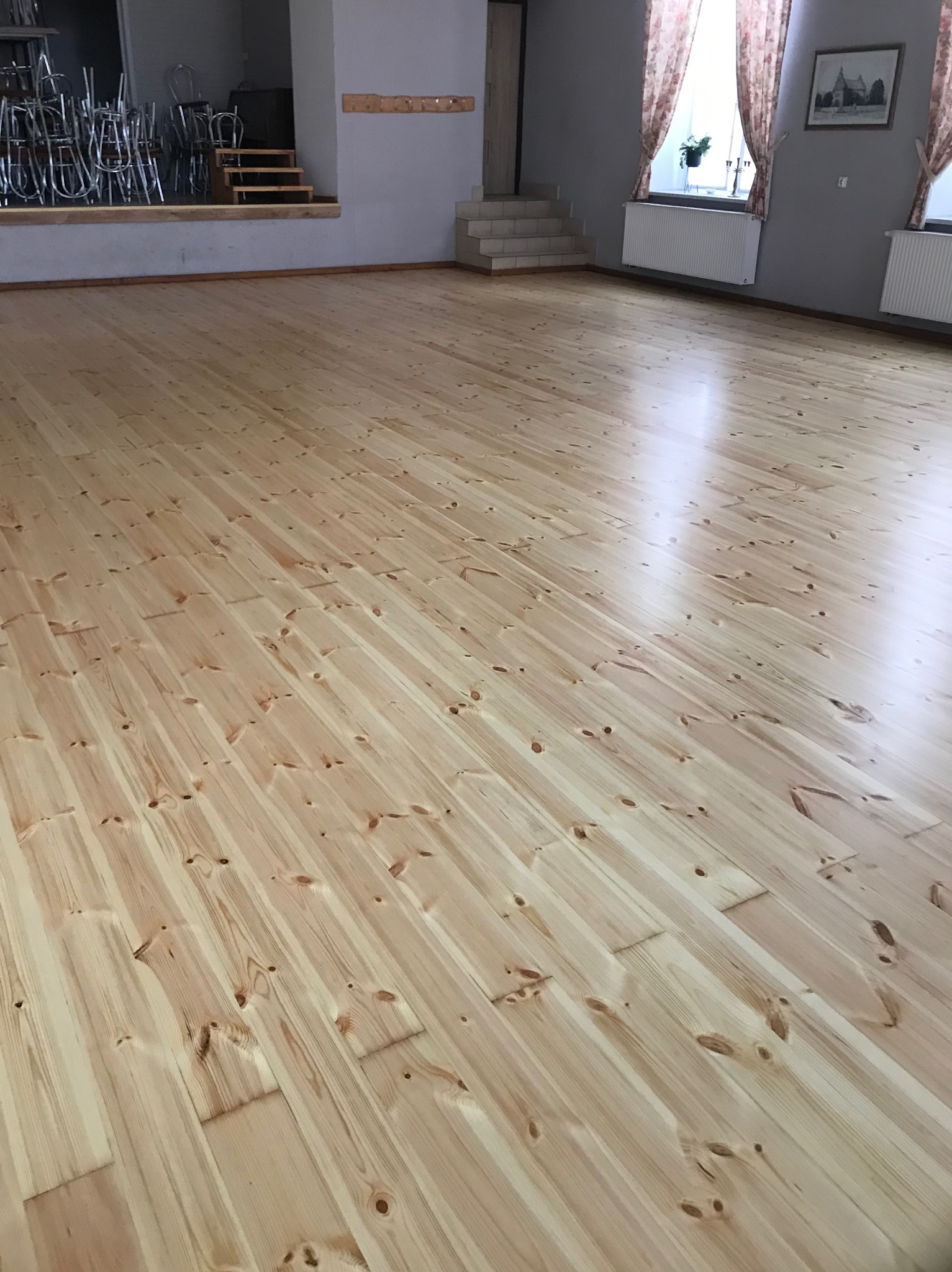 Jasna, drewniana podłoga z widocznymi słojami, po renowacji w dużej sali z szarymi ścianami, grzejnikami i oknami z zasłonami. Na podwyższeniu w tle widoczne krzesła.