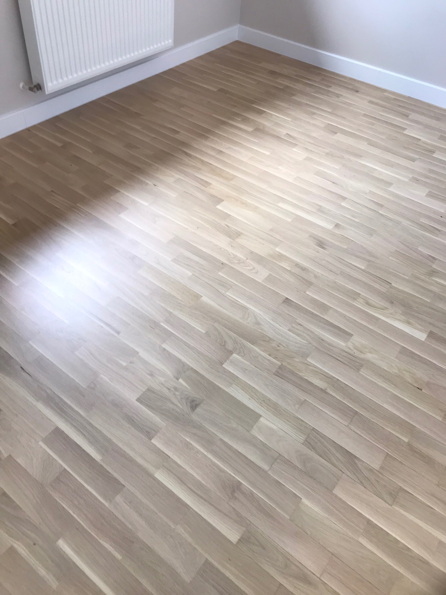 Jasna, wycyklinowana podłoga parkietowa ułożona w jodełkę, widoczny fragment grzejnika i białe listwy przypodłogowe, pomieszczenie po remoncie.
