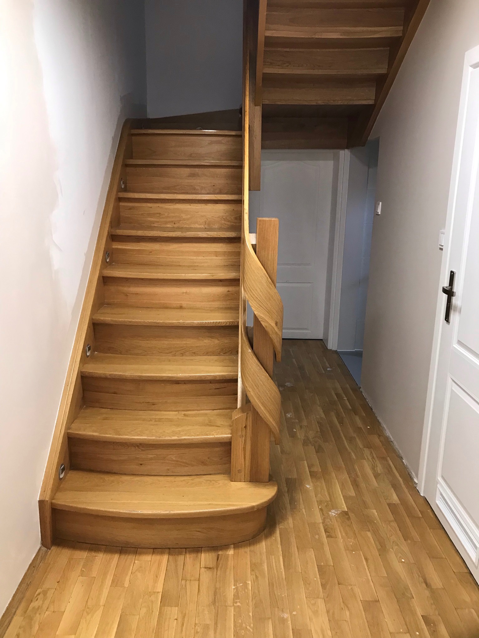 Drewniane schody z oświetleniem punktowym na każdym stopniu, z ozdobną, spiralną balustradą, prowadzące na piętro. Parkiet na podłodze dopasowany do koloru schodów.