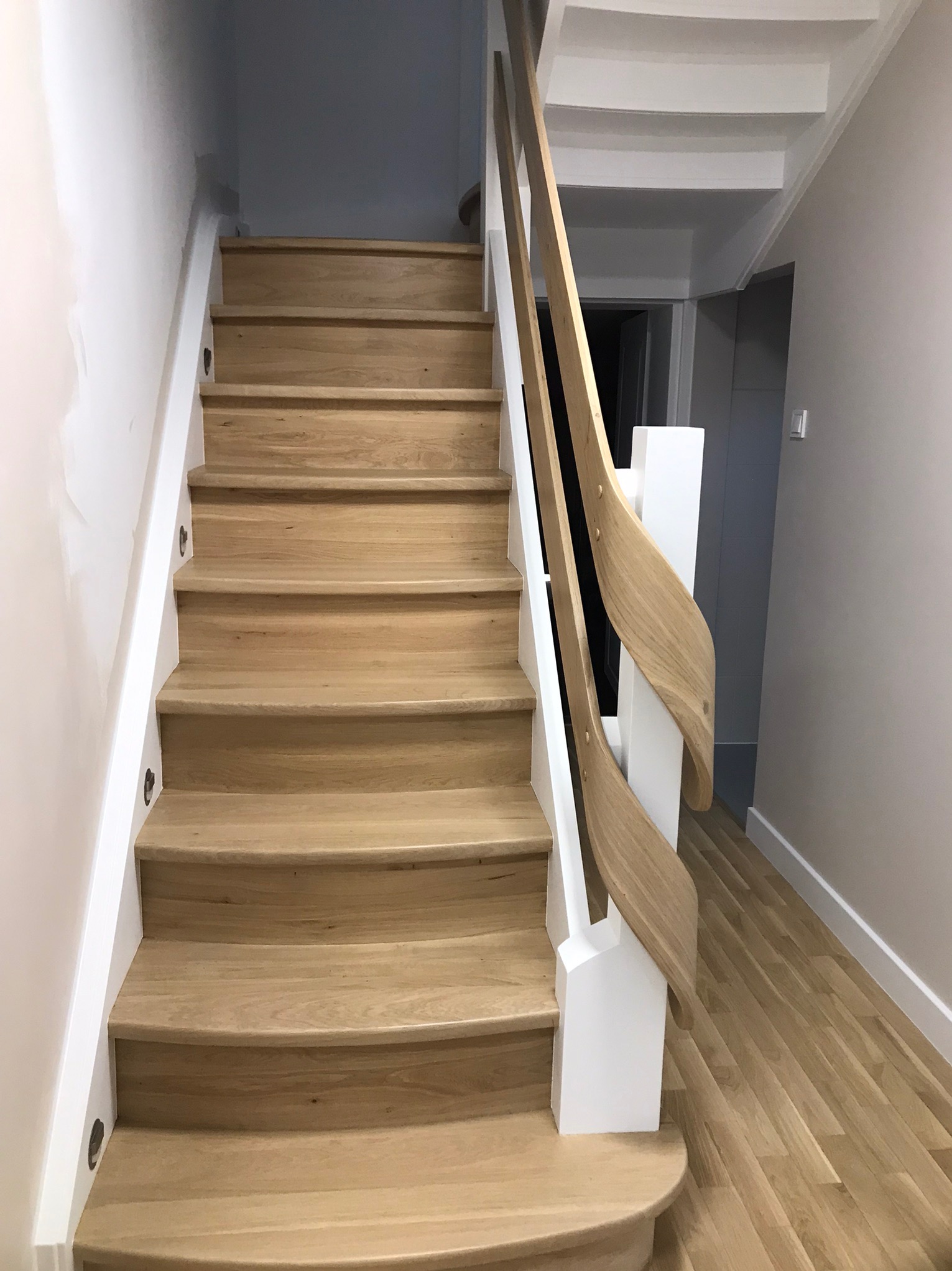 Jasne drewniane schody z białymi balustradami o zakrzywionym kształcie, oświetlenie punktowe na ścianie, widok z dołu na górę.