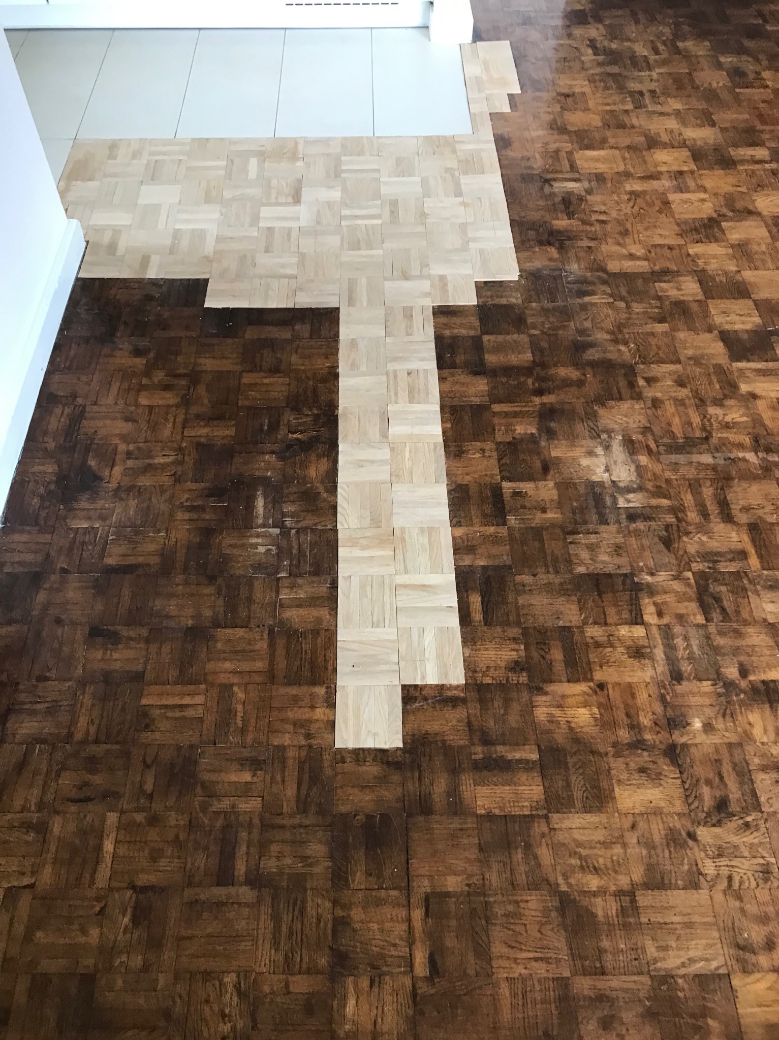 Fragment podłogi parkietowej w trakcie renowacji, widoczny kontrast między wycyklinowanym jasnym fragmentem w kształcie litery T a ciemniejszą, starą powierzchnią parkietu, w tle białe płytki...