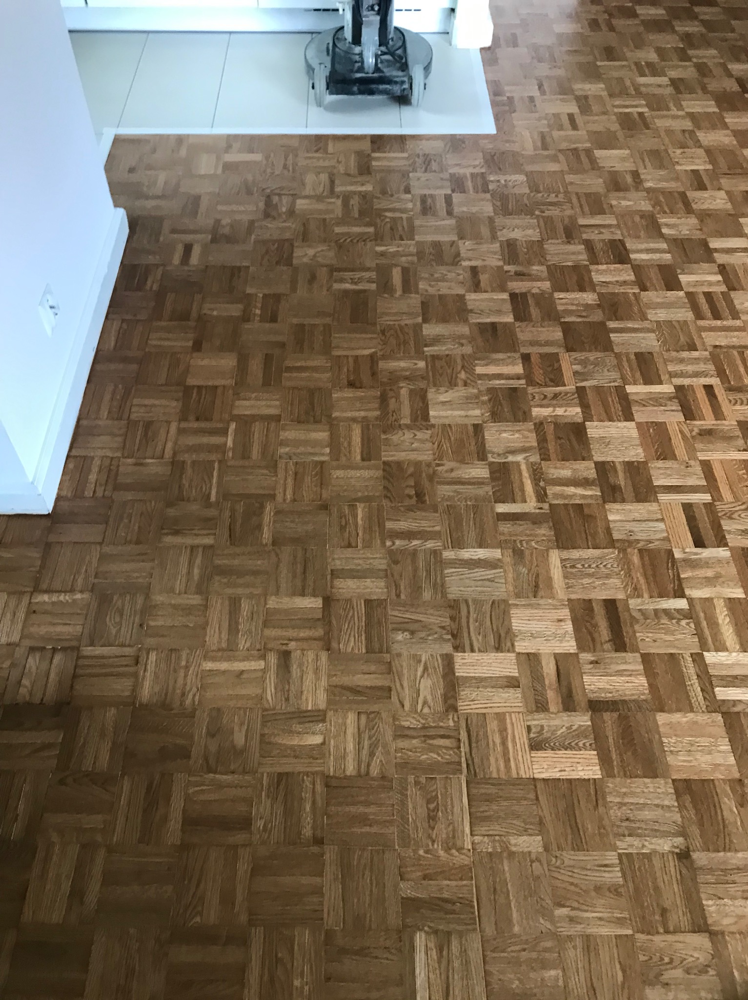 Parkiet ułożony w kwadraty, częściowo odnowiony maszyną do renowacji podłóg, widoczna biała listwa przypodłogowa i fragment białej podłogi.