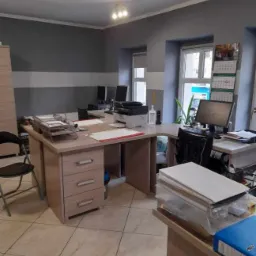 Wnętrze biura z biurkami, komputerami i dokumentami, widok na stanowiska pracy z krzesłami i kalendarzem na ścianie, oświetlenie sufitowe.