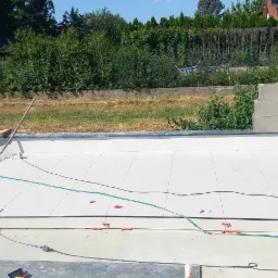 Układanie jasnych płytek na tarasie zewnętrznym, widoczne narzędzia glazurnicze, poziomica i elementy mocujące, w tle zielona roślinność i fragment ogrodzenia.