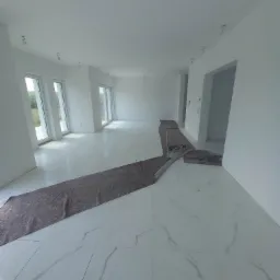 Wnętrze nowo wykończonego, przestronnego pokoju z białymi ścianami i marmurową podłogą zabezpieczoną brązową folią ochronną przed dalszymi pracami.