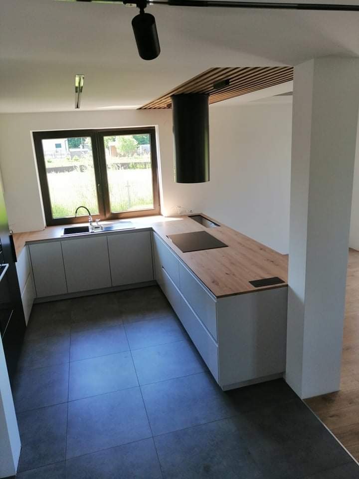 Nowoczesna kuchnia z szarymi szafkami, drewnianym blatem i czarnym okapem cylindrycznym oraz drewnianą listwą na suficie, widok na zewnątrz przez okno.