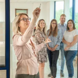 Kobieta w okularach pisze markerem po szklanej tablicy, za nią grupa osób obserwuje prezentację.