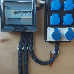 Szara skrzynka bezpiecznikowa z widocznymi bezpiecznikami Eaton i Legrand, oraz czarna skrzynka z sześcioma gniazdami ABL (cztery niebieskie i dwa białe), połączone szarymi, karbowanymi przewodami...