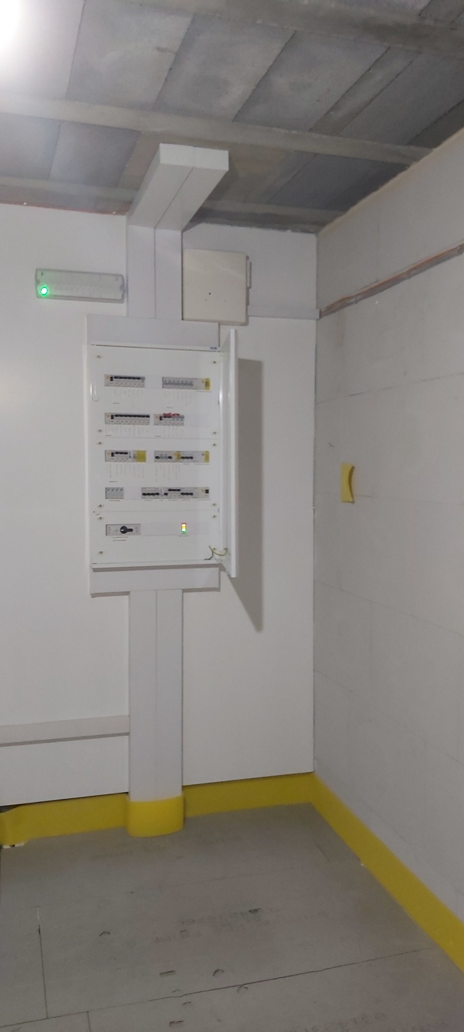 Otwarta skrzynka bezpiecznikowa z białym wspornikiem na pomalowanej na biało ścianie, widoczne elementy instalacji elektrycznej, podłoga zabezpieczona żółtą folią malarską.