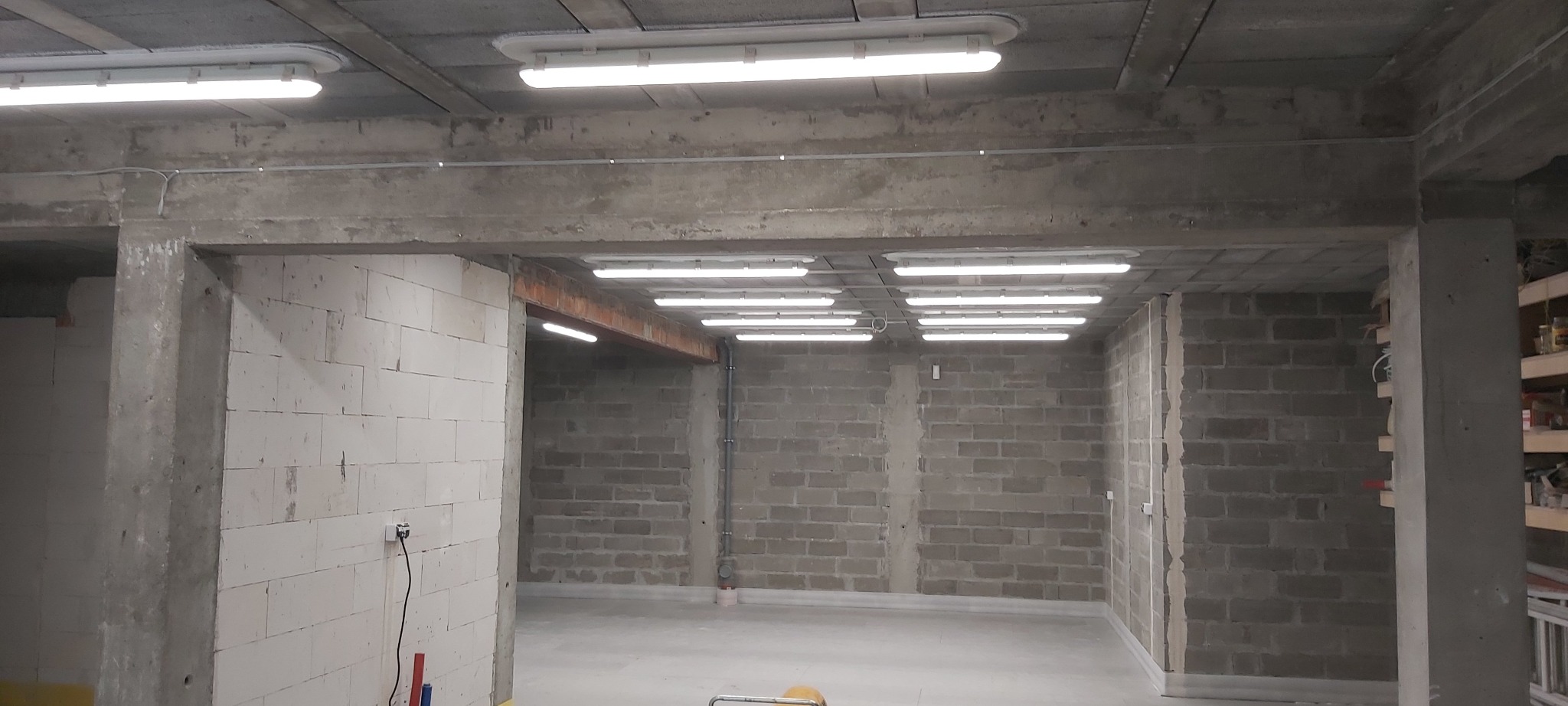 Surowe wnętrze w trakcie remontu: ściany z bloczków betonowych i betonu komórkowego, sufit z widocznymi lampami jarzeniowymi, widoczne instalacje elektryczne, stan deweloperski.