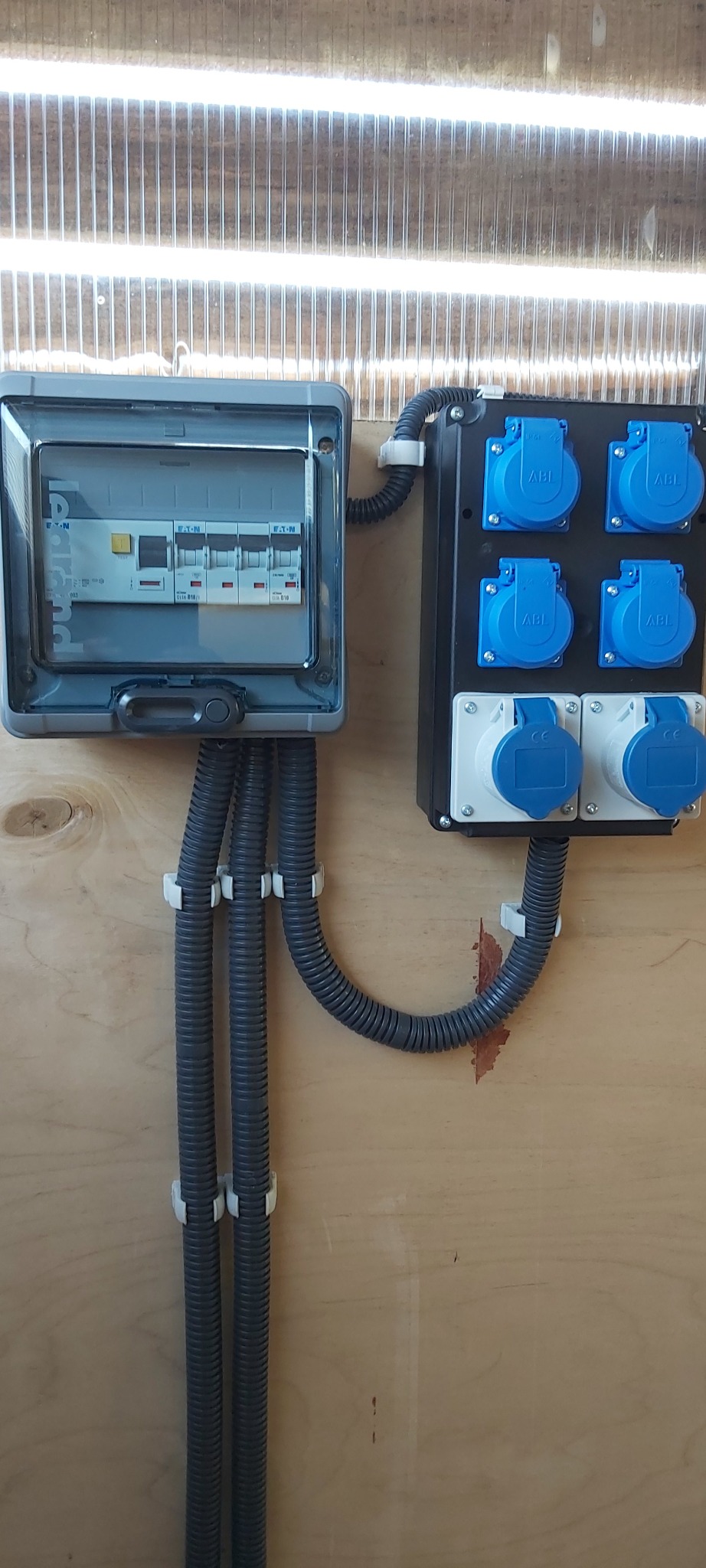 Szara skrzynka bezpiecznikowa z widocznymi bezpiecznikami Eaton i Legrand, oraz czarna skrzynka z sześcioma gniazdami ABL (cztery niebieskie i dwa białe), połączone szarymi, karbowanymi przewodami...