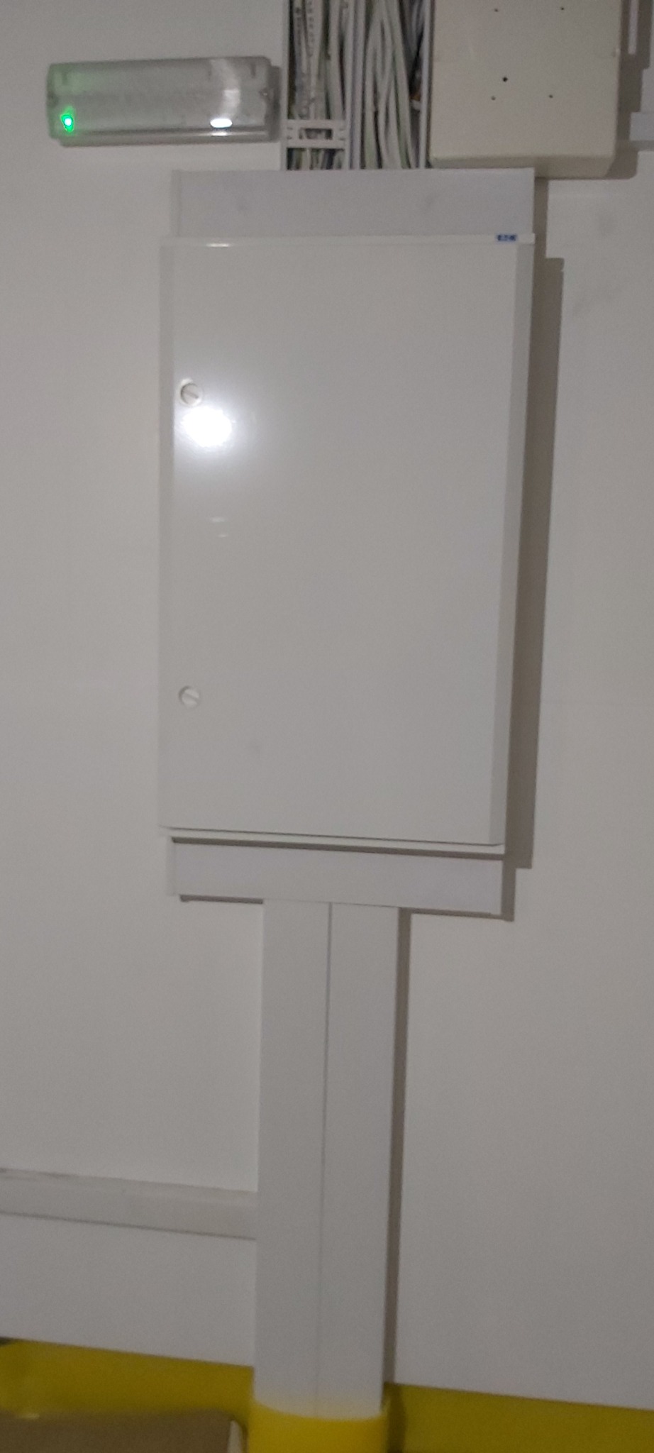 Biała skrzynka elektryczna z widocznym okablowaniem na ścianie, z zieloną lampką kontrolną u góry.