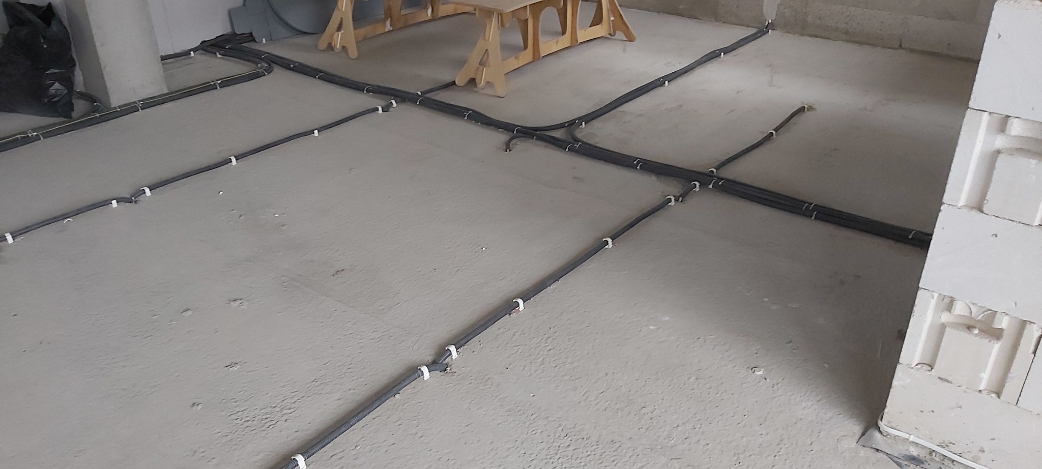 Szare rury elektryczne w peszlu, przymocowane białymi uchwytami do surowej podłogi w trakcie remontu, widoczne bloczki betonowe i drewniany kozioł.