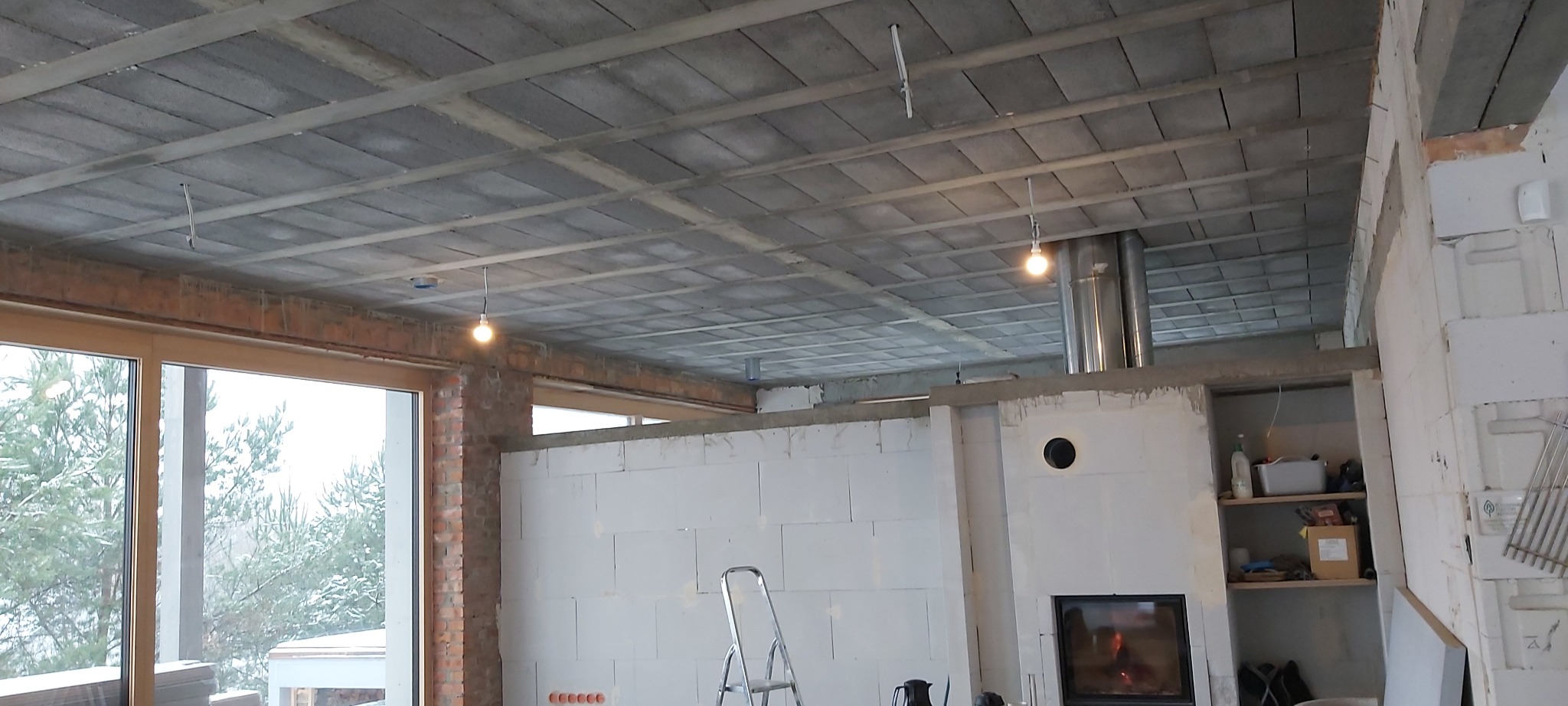 Surowy stan wnętrza: widoczny strop z betonowych płyt, niedokończona ściana z białych bloczków, komin i przygotowane miejsce na kominek, surowe okno z widokiem na zimowy krajobraz.