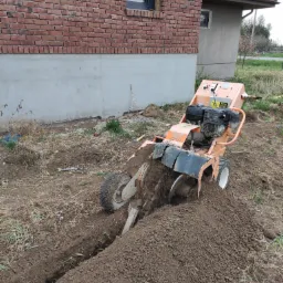 Pomarańczowa glebogryzarka spalinowa w trakcie kopania wąskiego rowu w ziemi przy budynku z czerwonej cegły.