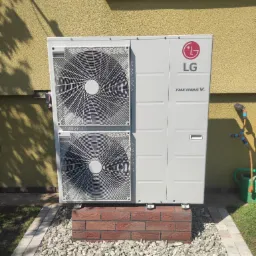 Zewnętrzna jednostka pompy ciepła LG Therma V, umieszczona na cokole z czerwonej cegły, otoczona drobnymi kamieniami, z żółtą elewacją budynku w tle.