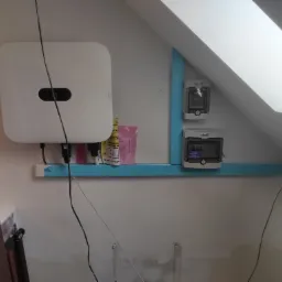 Biały inwerter fotowoltaiczny zamontowany na ścianie pod skosem dachu, obok zabezpieczenia w niebieskiej obudowie i opakowania po akcesoriach montażowych.