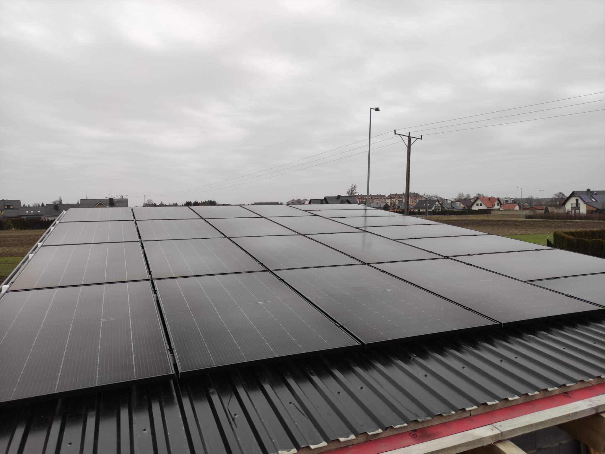 Instalacja fotowoltaiczna na dachu z blachy trapezowej, widok na okoliczne domy i słup energetyczny w pochmurny dzień.