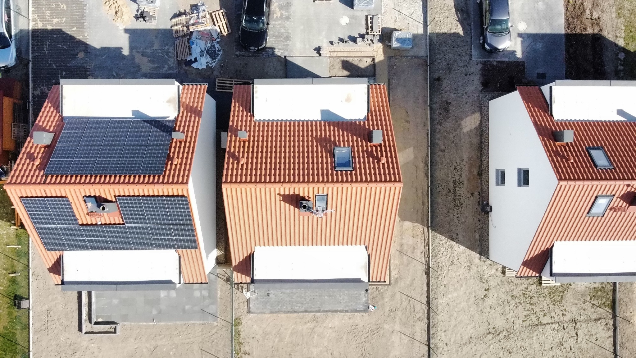 Widok z lotu ptaka na trzy domy jednorodzinne z brązową dachówką, jeden z zainstalowanymi panelami fotowoltaicznymi na dachu, zaparkowane samochody w pobliżu.