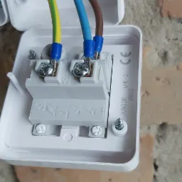 Otwarta puszka elektryczna z podłączonymi przewodami (żółto-zielony, niebieski, brązowy) zabezpieczonymi niebieskimi tulejkami, widoczne śruby mocujące na tle ceglanej ściany.