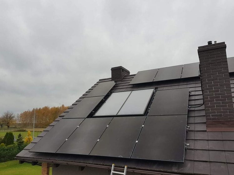Czarne panele słoneczne fotowoltaiczne zamontowane na brązowym dachu skośnym domu jednorodzinnego z oknem dachowym i ceglanym kominem, widoczne kable i drabina oparta o rynnę.