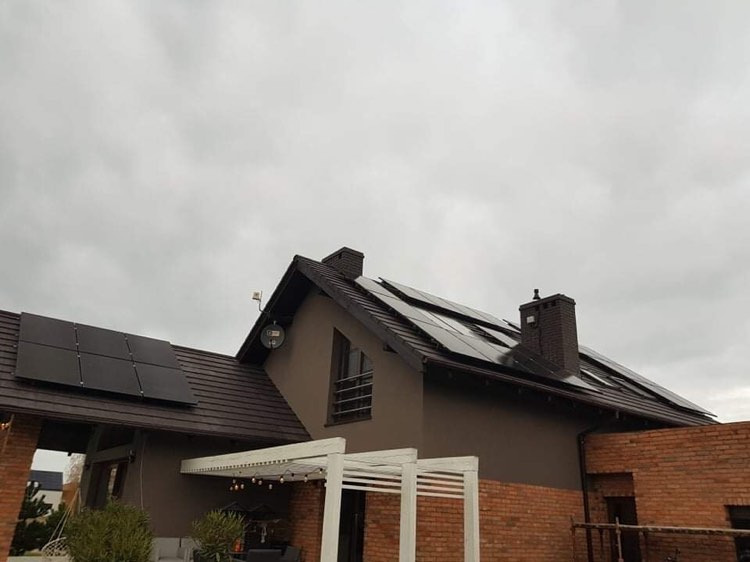 Czarny panel słoneczny na brązowym dachu domu jednorodzinnego z ceglaną elewacją, widoczne elementy pergoli i pochmurne niebo.