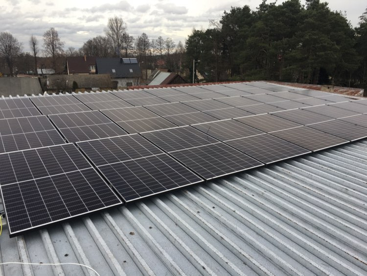 Rząd paneli fotowoltaicznych na blaszanym dachu budynku gospodarczego, widok z góry przy pochmurnej pogodzie, w tle zabudowa wiejska i drzewa.