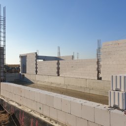 Makerbud - Wznoszenie ścian fundamentowych z bloczków betonowych, widoczna izolacja przeciwwilgociowa i zbrojenie pionowe, w tle fragment konstrukcji domu w stanie surowym otwartym.