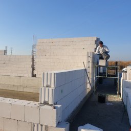 Makerbud - Murarz na rusztowaniu w trakcie wznoszenia ściany z białych bloczków, widoczne zbrojenia pionowe, zapas bloczków obok, w tle pole kukurydzy i słupy wysokiego napięcia.