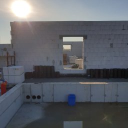 Makerbud - Wznoszone ściany domu z betonu komórkowego na fundamencie, widoczne otwory okienne i drzwiowe, palety z bloczkami oraz czarne rolki izolacyjne w tle, wczesna faza budowy.