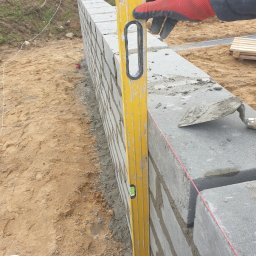 Makerbud - Wznoszenie ściany z szarych bloczków na zaprawie, kontrolowane poziomicą przez pracownika w czerwonej rękawicy roboczej, widoczna kielnia i napięty sznurek murarski.