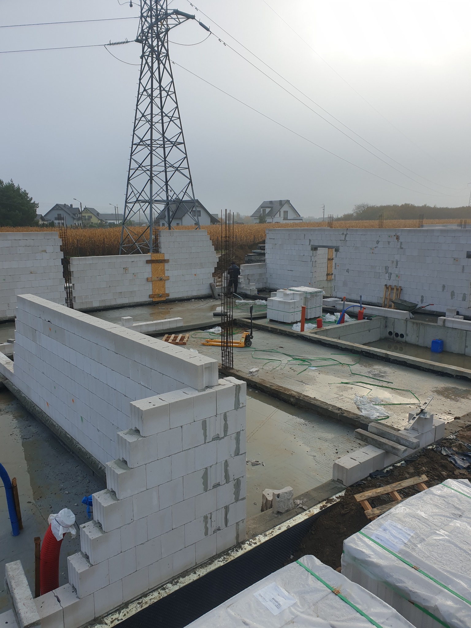 Wznoszenie ścian domu z bloczków betonu komórkowego, widoczne zbrojenia, paleta z bloczkami w folii, w tle słup wysokiego napięcia i zabudowa jednorodzinna.