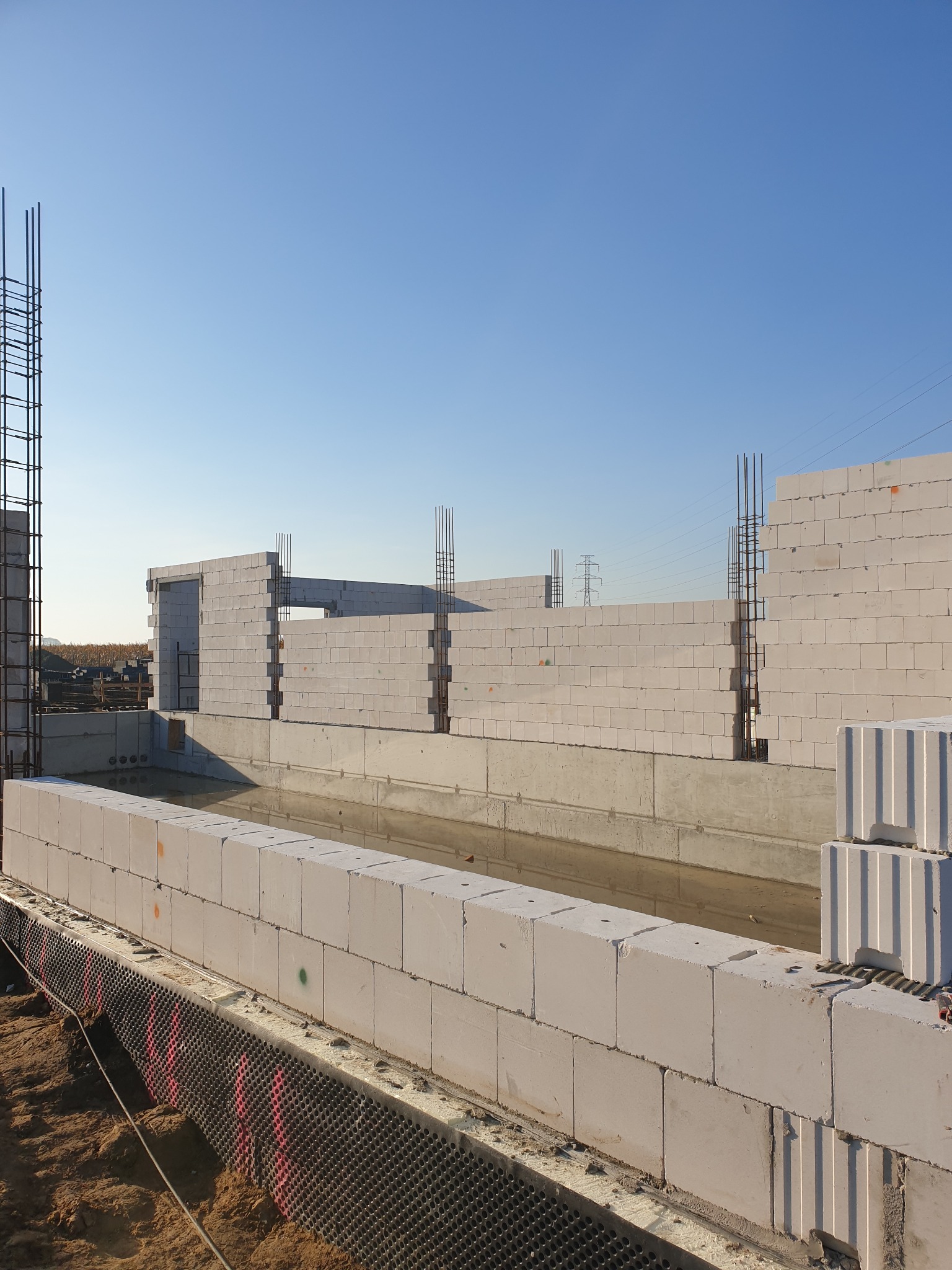 Wznoszenie ścian fundamentowych z bloczków betonowych, widoczna izolacja przeciwwilgociowa i zbrojenie pionowe, w tle fragment konstrukcji domu w stanie surowym otwartym.