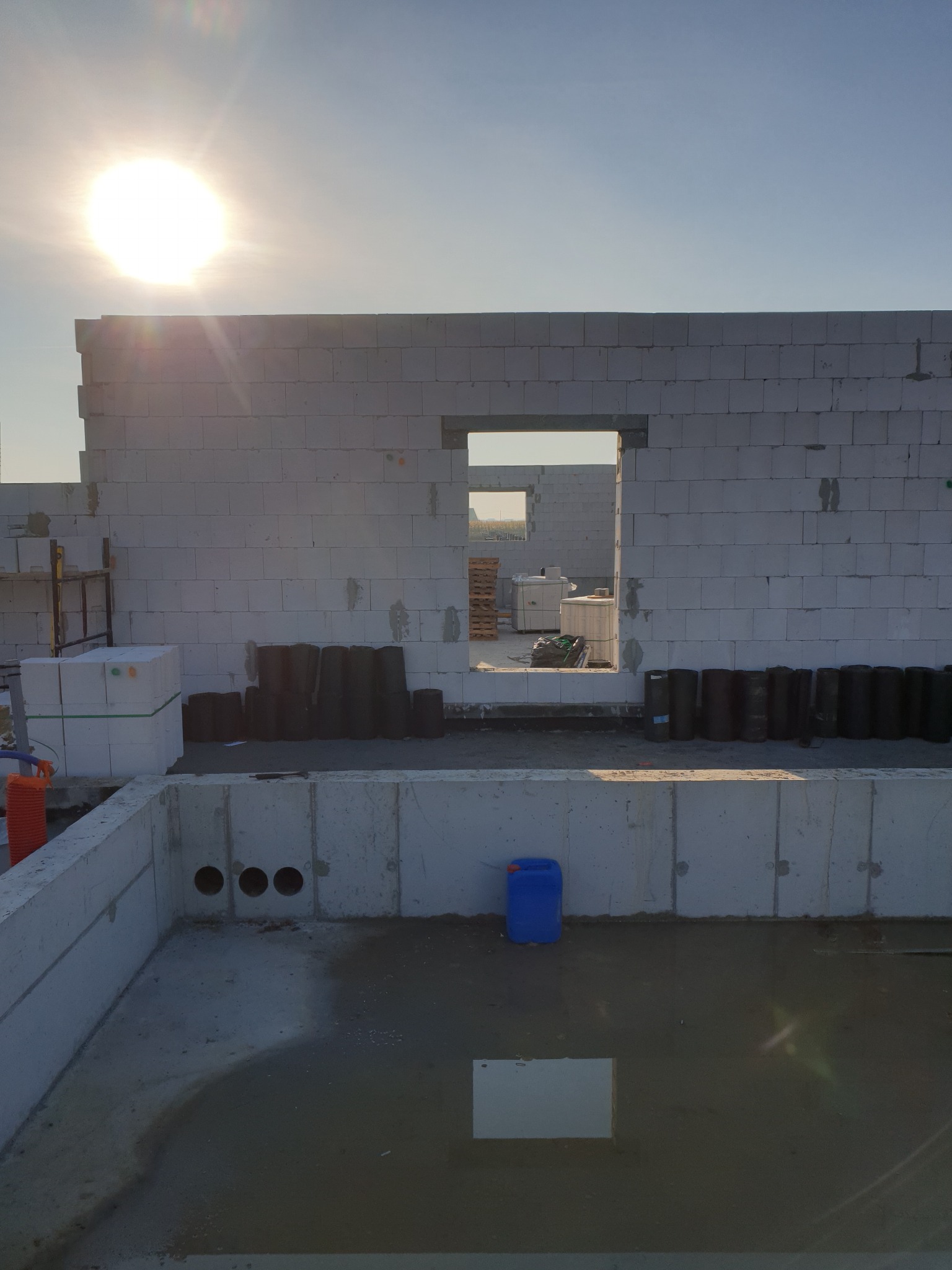 Wznoszone ściany domu z betonu komórkowego na fundamencie, widoczne otwory okienne i drzwiowe, palety z bloczkami oraz czarne rolki izolacyjne w tle, wczesna faza budowy.