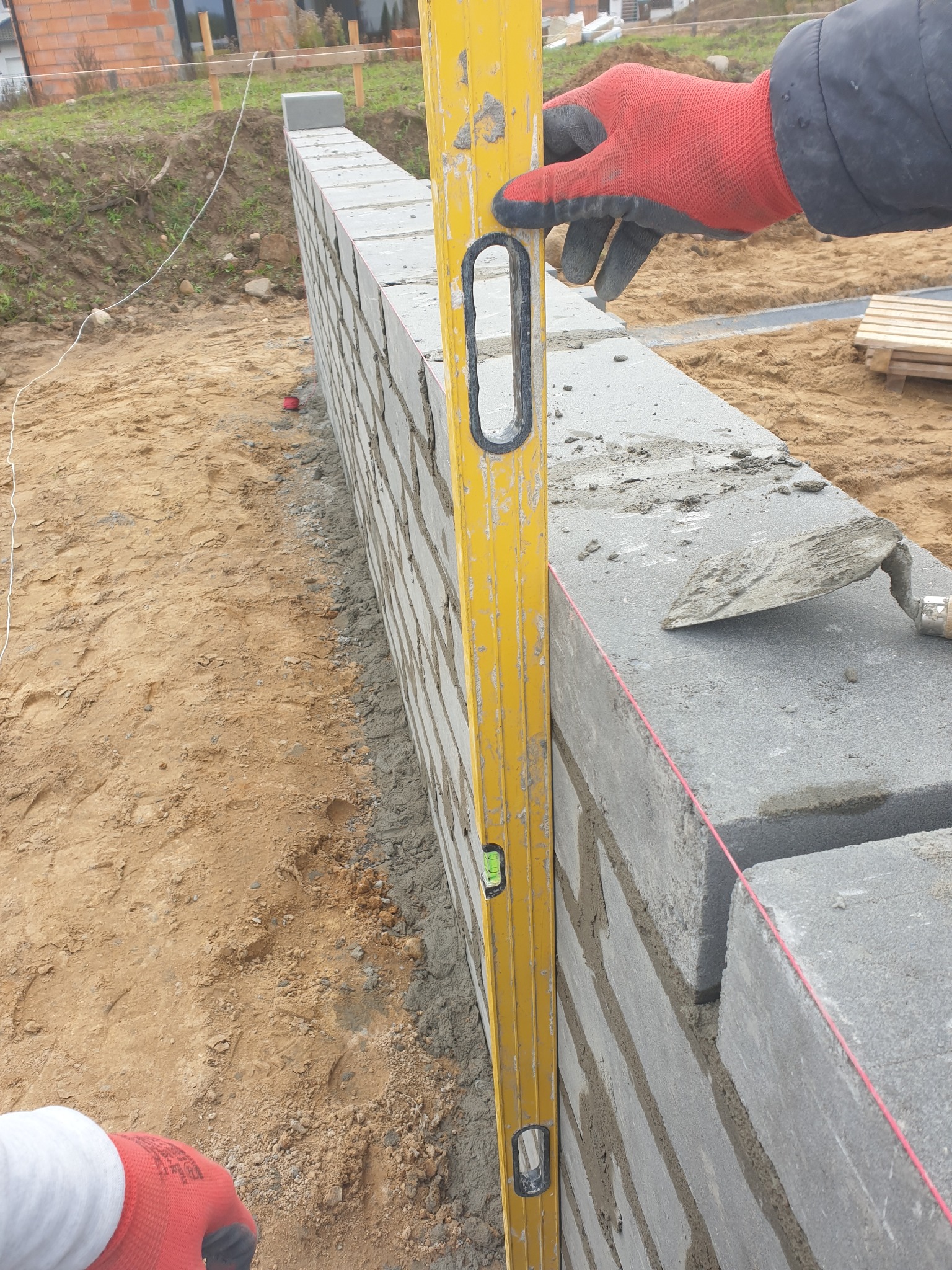 Wznoszenie ściany z szarych bloczków na zaprawie, kontrolowane poziomicą przez pracownika w czerwonej rękawicy roboczej, widoczna kielnia i napięty sznurek murarski.