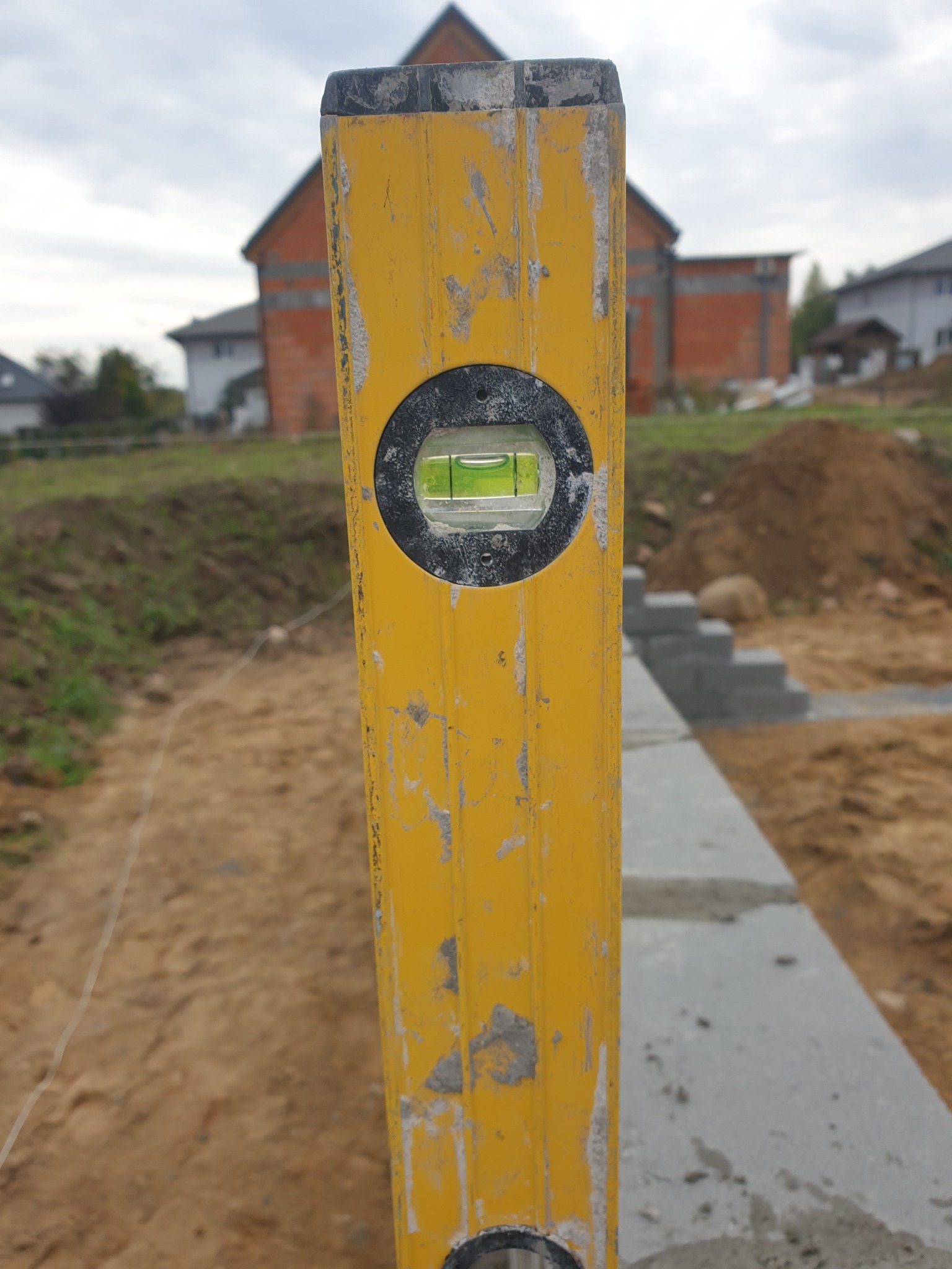 Żółta poziomica w pionie, ustawiona na fundamencie z bloczków betonowych, w tle budowa domu z czerwonej cegły i inne budynki.